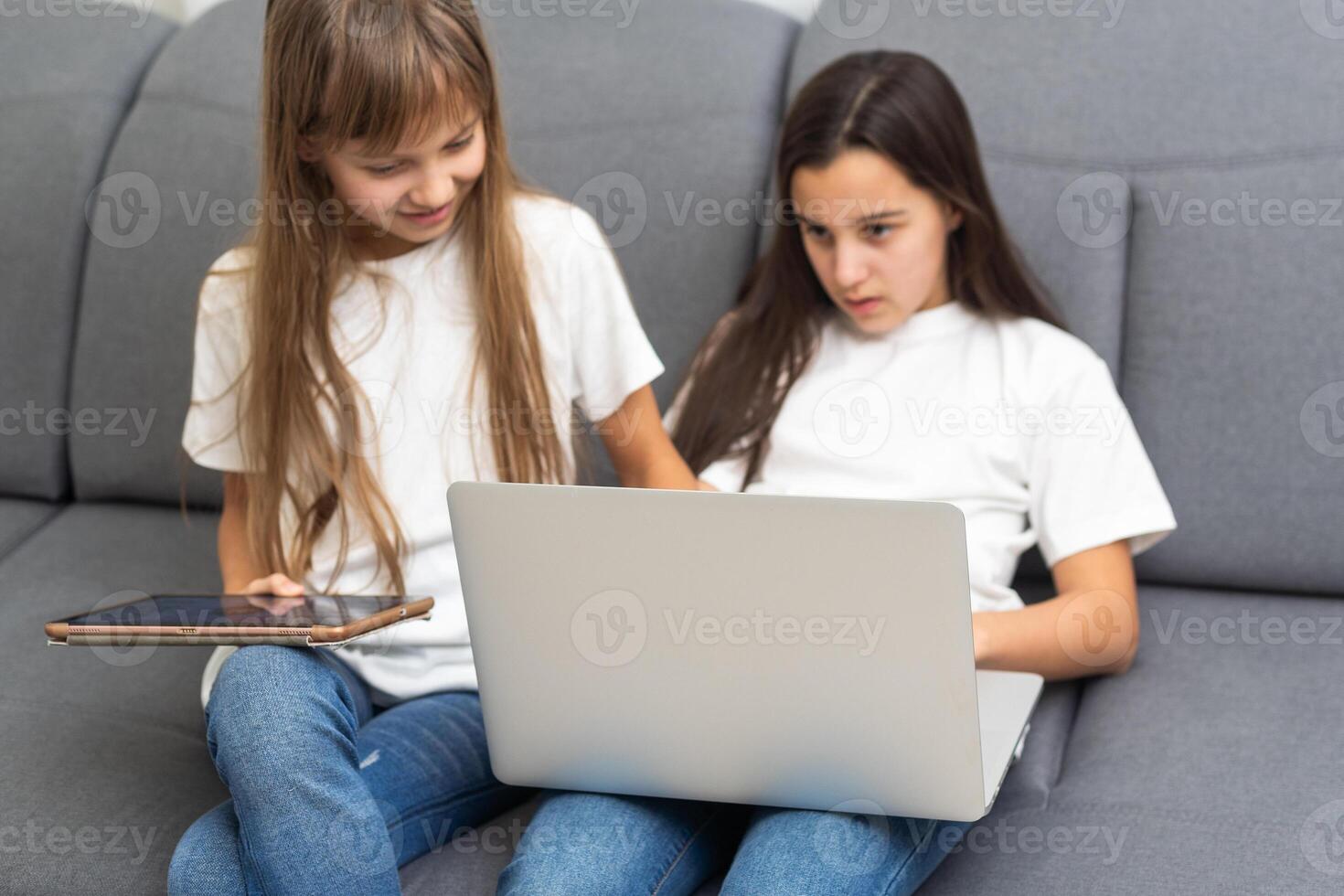Cute little girl and older sister playing together smiling and having bonding time using a laptop on couch at home. Happy family Siblings relationship and digital technology lifestyle concept. photo