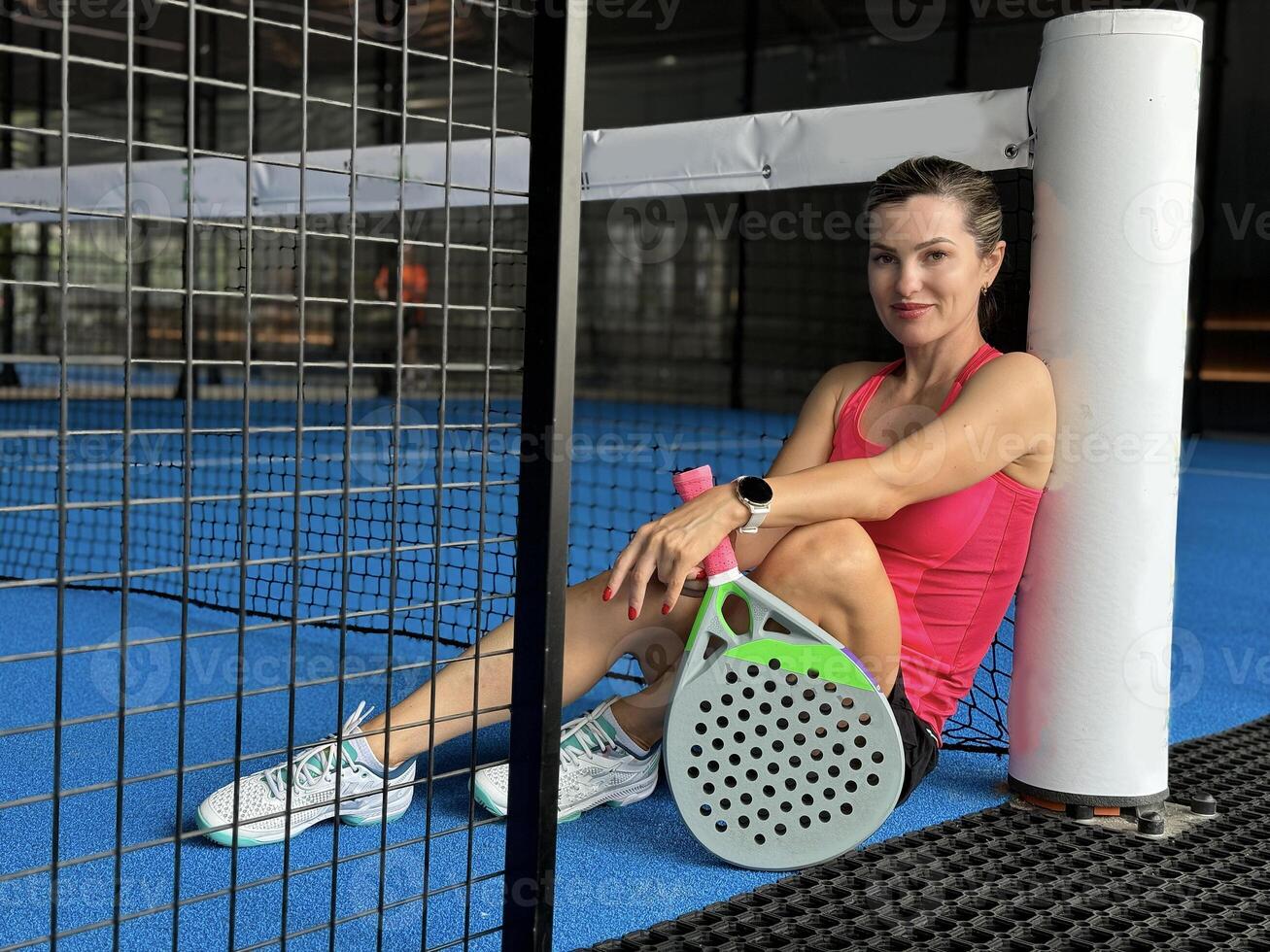 Sportive young girl with racquet at net in padel court, smiling and looking at camera. photo