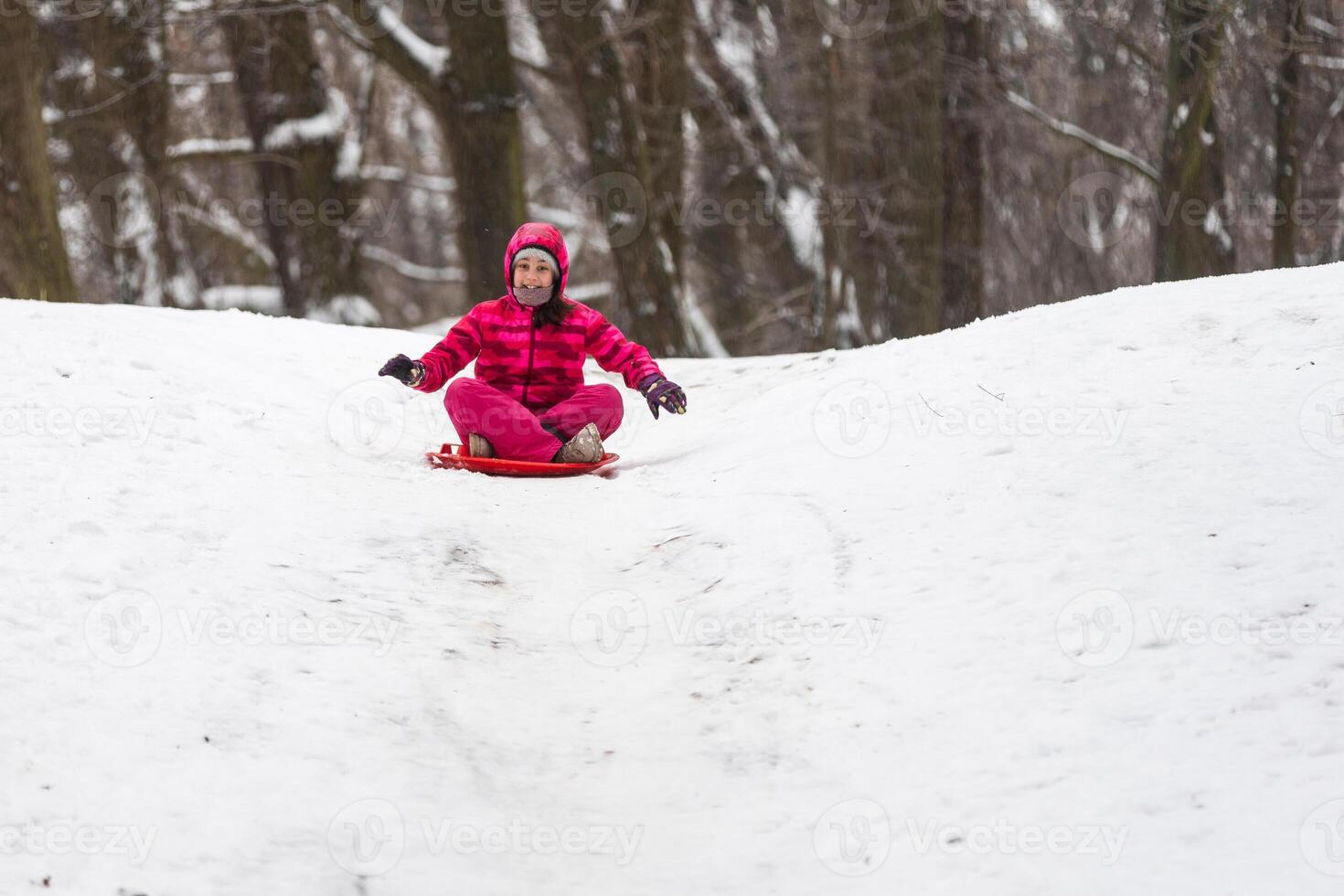 childhood, sledging and season concept - happy little girl sliding down on sled outdoors in winter photo