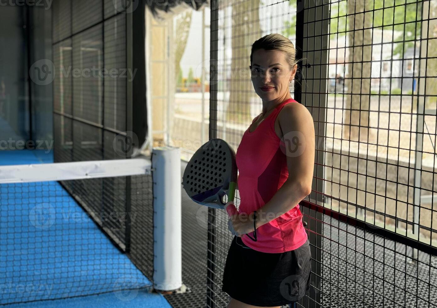 Sportive young girl with racquet at net in padel court, smiling and looking at camera. photo