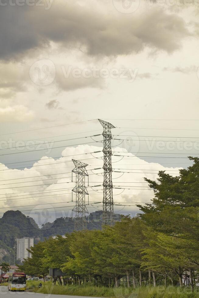 Tall power transmission tower with multiple cables stretch across the sky, set against a cloudy backdrop. Lush greenery and trees fill the foreground, contrasting with the industrial structures above. photo