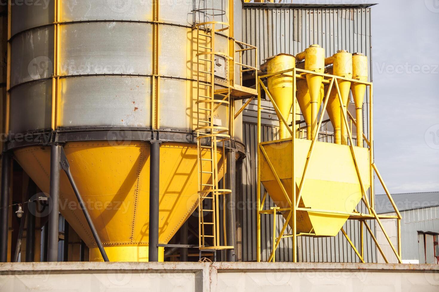 large grain elevator with multiple cylindrical metal silos, structural elements, and a complex system of pipes. It stands against a blue sky, with a field in the foreground after harvest photo