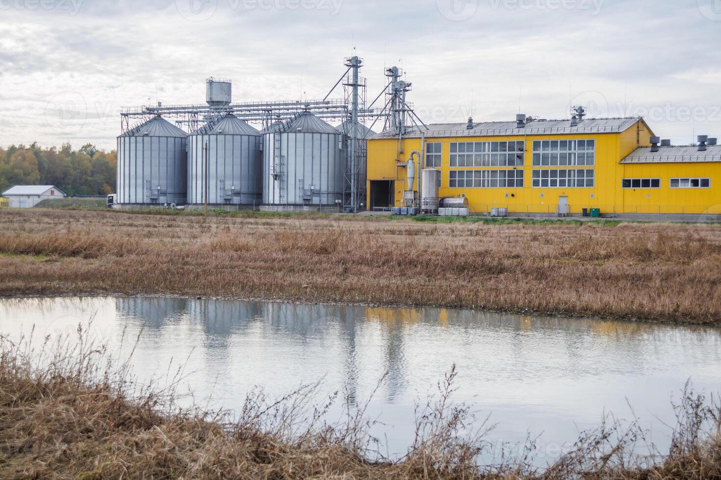 large grain elevator with multiple cylindrical metal silos, structural elements, and a complex system of pipes. It stands against a blue sky, with a field in the foreground after harvest photo