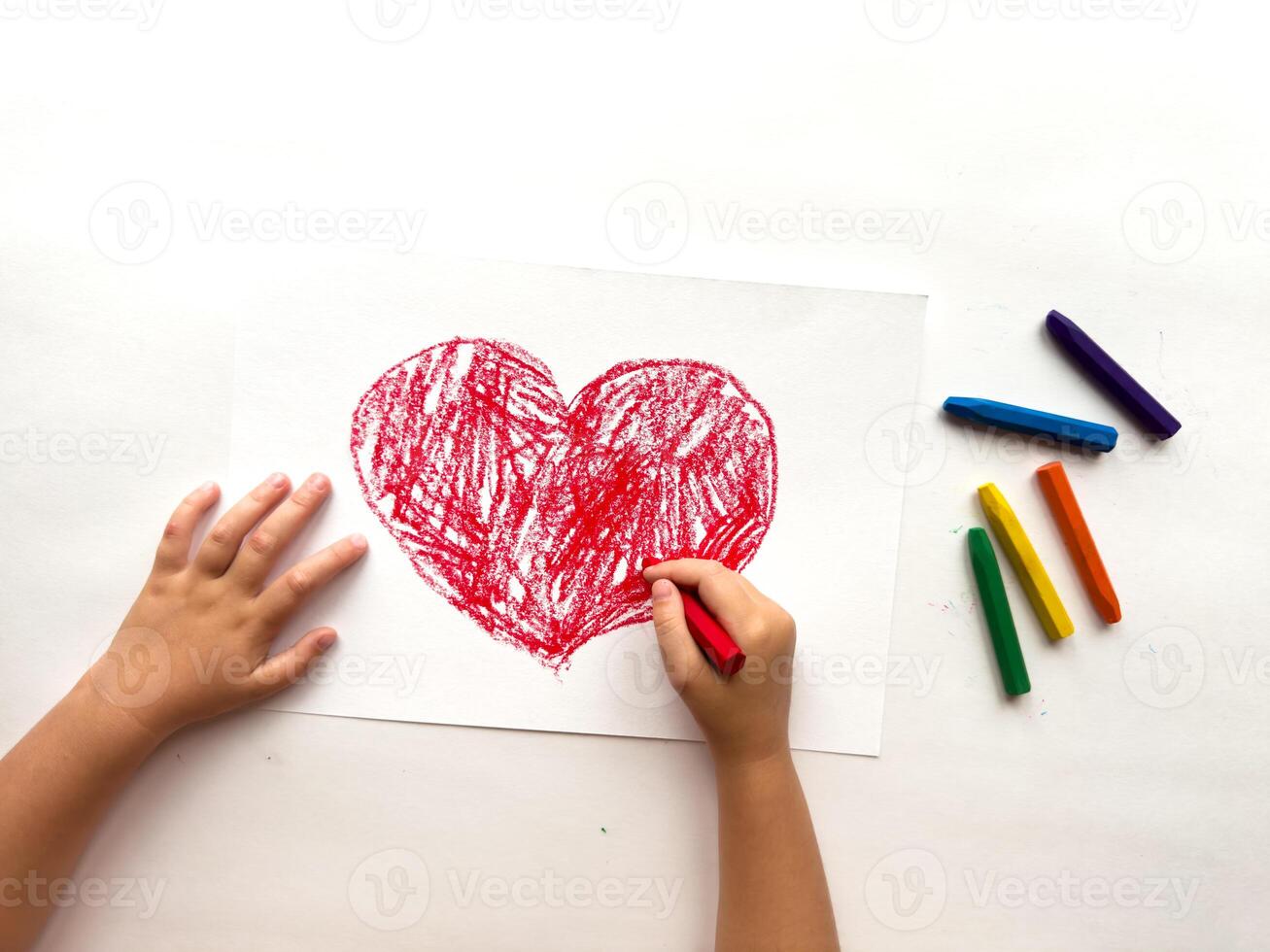 Child drawing a red heart with crayons on white paper photo
