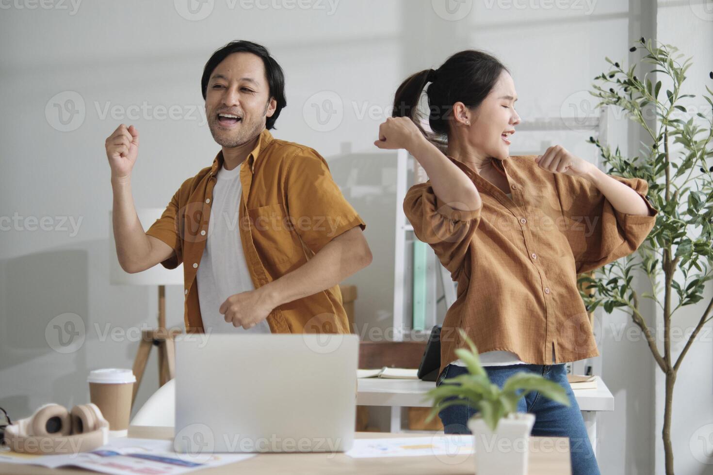Happy working in casual small business office. Two Asian startup company colleagues enjoy dancing and singing songs together, relaxing from freelance jobs, humor, and funny coworker team in workplace. photo