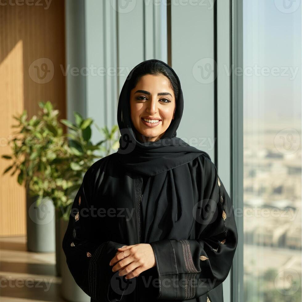 A friendly , candid portrait of a Saudi woman smiling confidently, dressed in traditional abaya and hijab, standing by a large window in a sleek, modern office photo