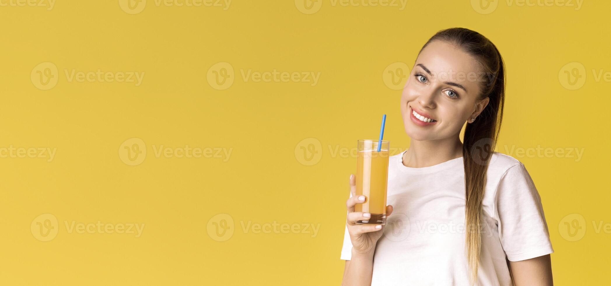Portrait of smiling young woman with glass of orange vitamin juice in hand, holding glass with straw, posing over yellow background, free space photo