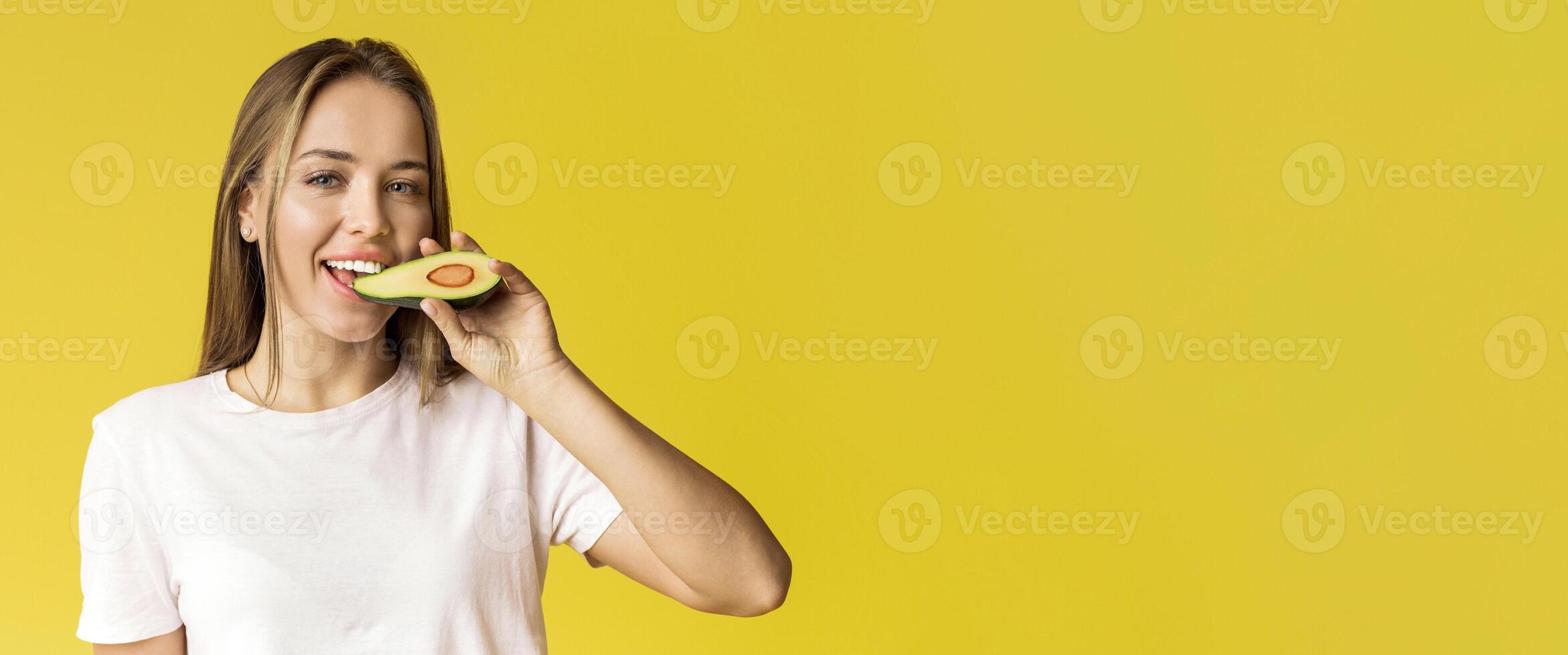 Healthy Diet. Happy Young Woman Biting Avocado Half And Smiling To Camera While Posing Over Yellow Studio Background With Empty Space photo