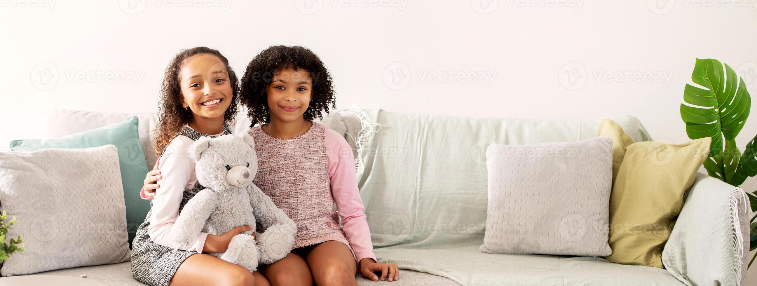 Two black girls with curly hair sit on a cozy sofa, one holding a soft teddy bear. They are smiling in a warm, inviting living room filled with colorful cushions. photo
