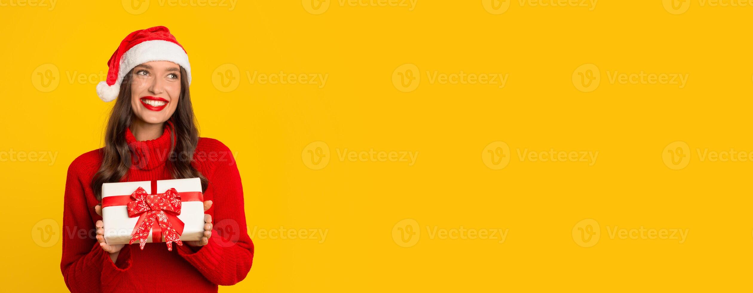 A smiling woman dressed in festive attire stands against a bright yellow background, happily holding a wrapped Christmas gift with a red bow, embodying the holiday spirit, copy space photo