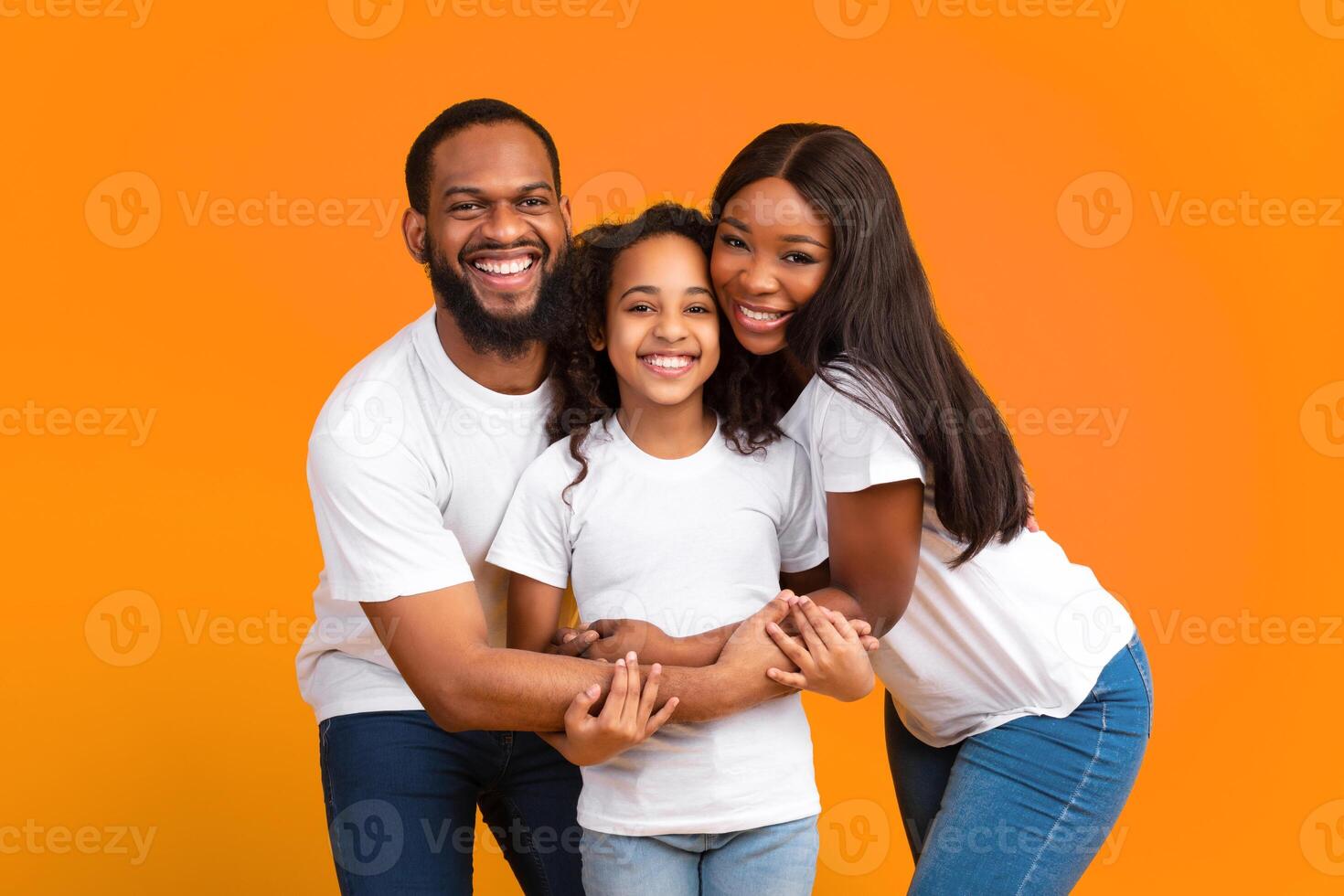 Love And Care Concept. Portrait of African American mum and dad embracing smiling daughter standing isolated on yellow studio background. Cheerful man and woman posing with girl and looking at camera photo