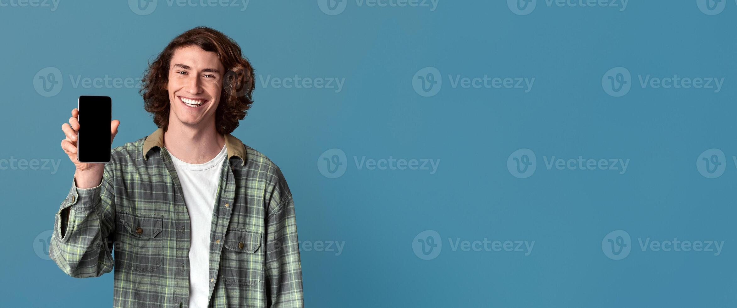 Smiling Caucasian guy holding smartphone with empty screen on turquoise background, space for your design photo