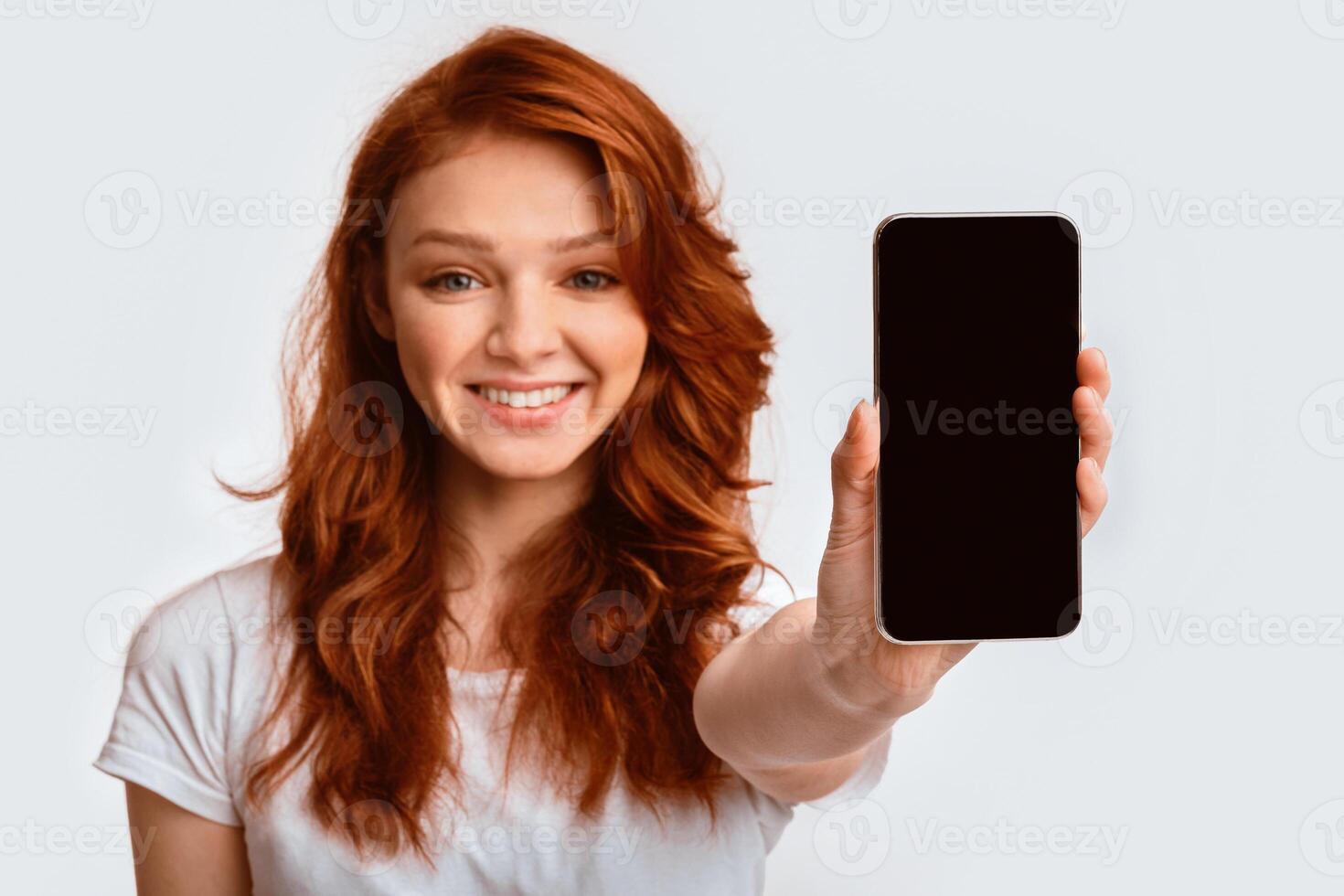Mobile App. Smiling Ginger-Haired Girl Showing Cellphone Blank Screen Recommending Application On White Background. Mockup, Studio Shot photo