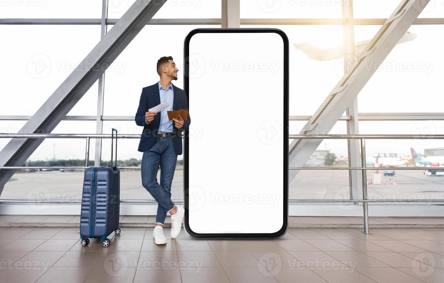 Middle eastern man in a suit stands casually near a large blank screen while holding his boarding pass and phone. He waits at the airport, glancing out the window at the planes. photo