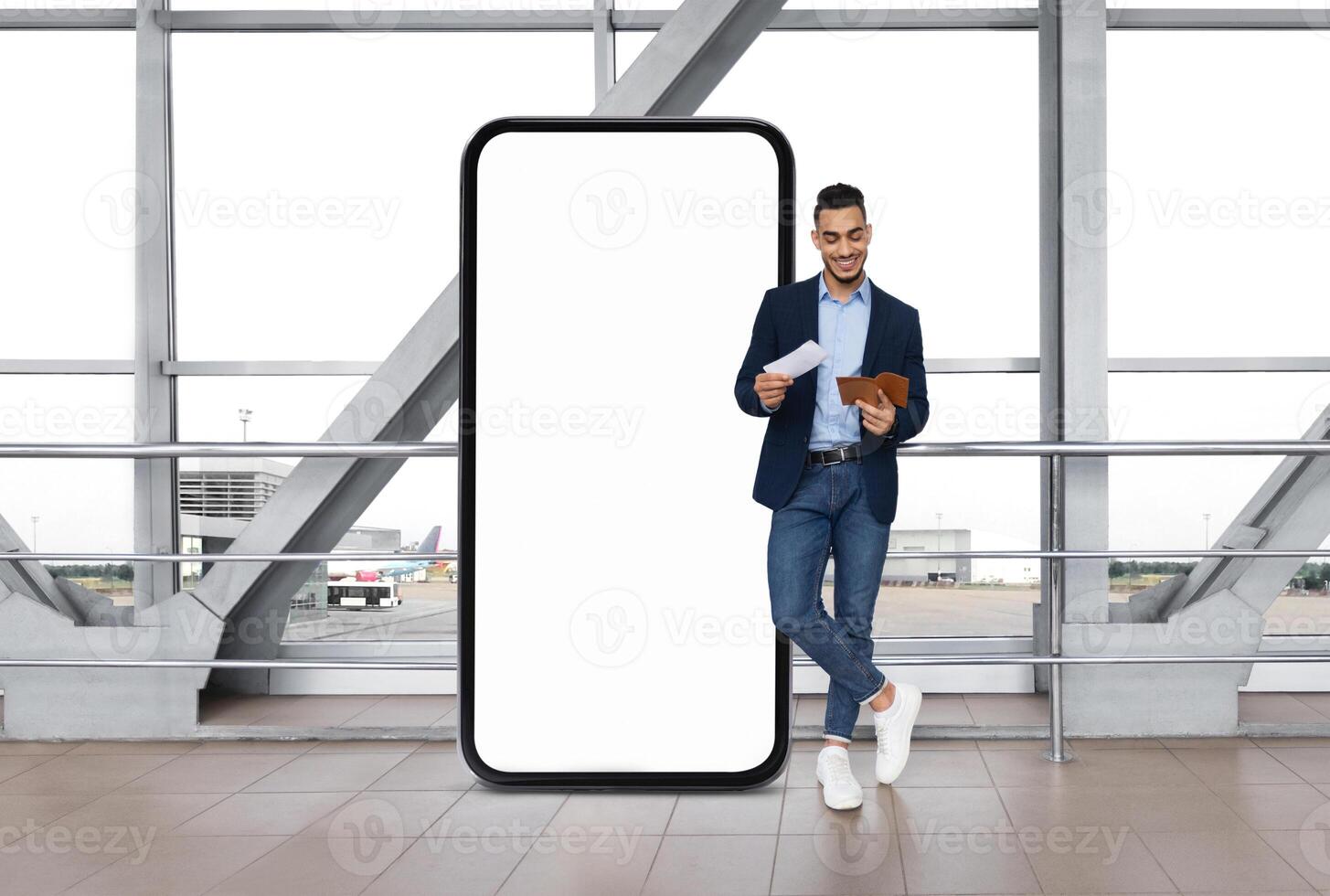 Arab man in a business suit stands against a large blank digital display at an airport terminal, looking at his tickets while holding a smartphone and a document, waiting for his flight. photo