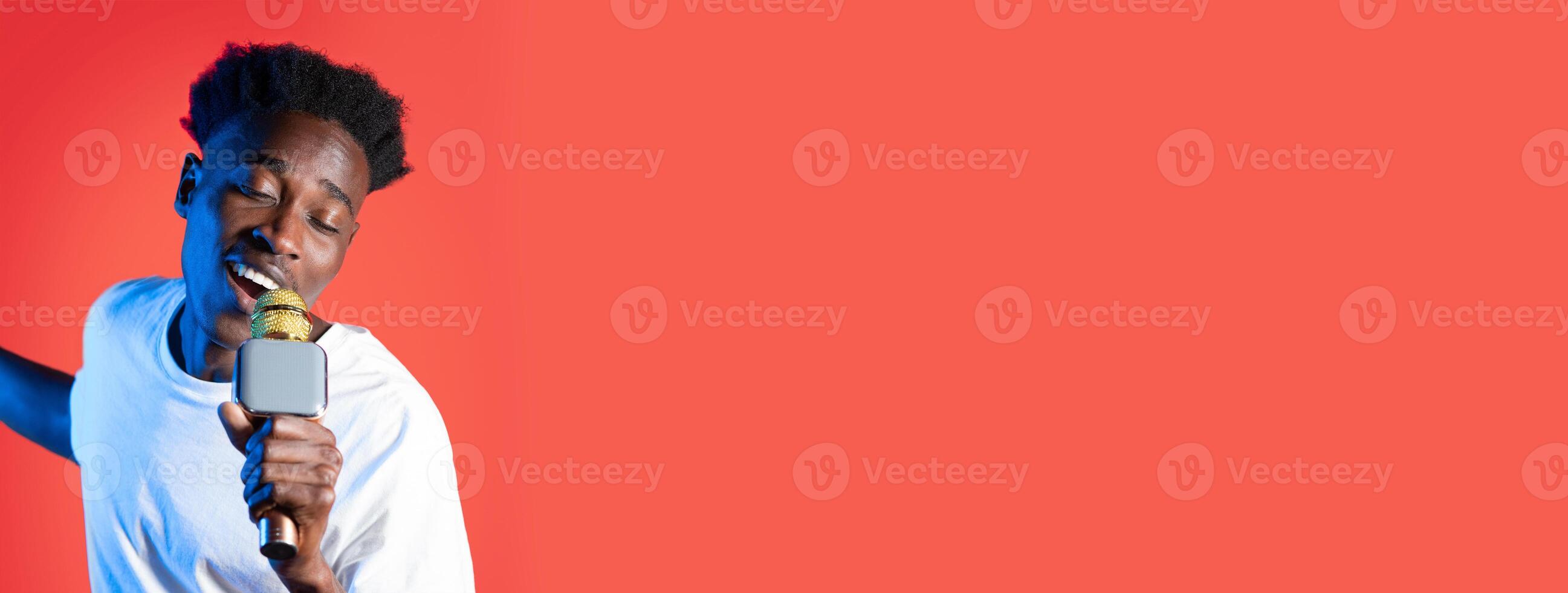 African American young man enjoys singing with a microphone in hand against a bright coral background. His expression reflects joy and enthusiasm for music and performance. photo