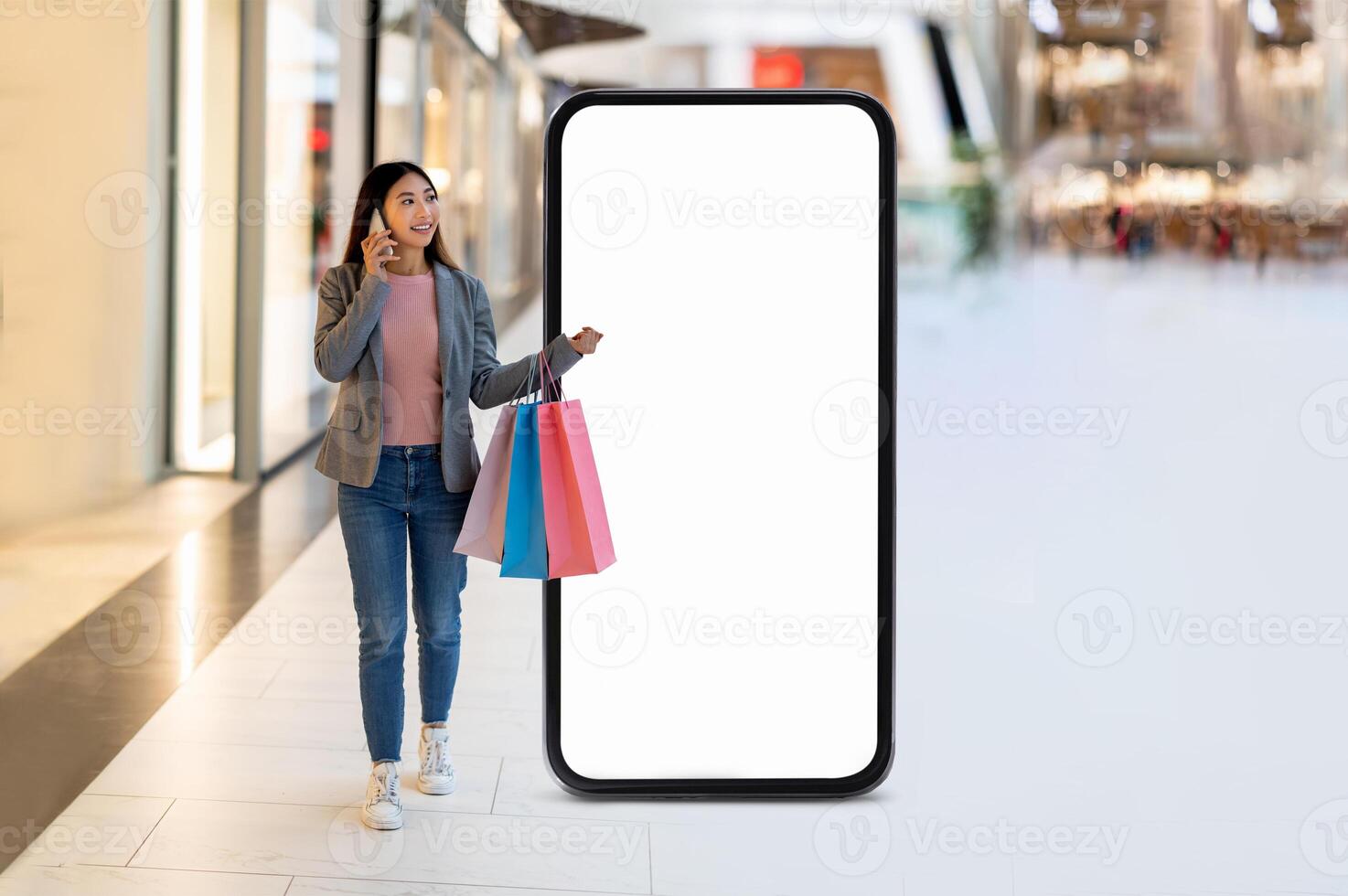 A young woman walks through a bustling shopping mall pass by huge phone with blank screen, chatting on her phone while carrying colorful shopping bags. photo