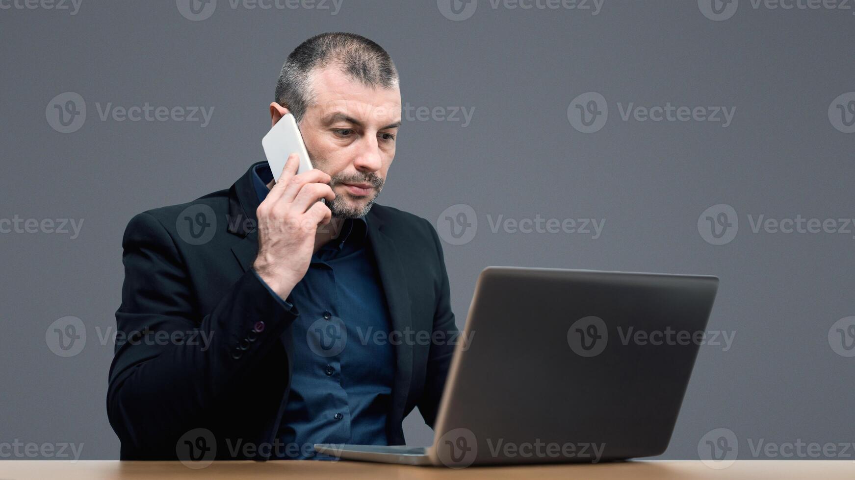 Distant Work. Mature Entrepreneur Talking On Cellphone Working On Laptop Sitting On Gray Studio Background photo
