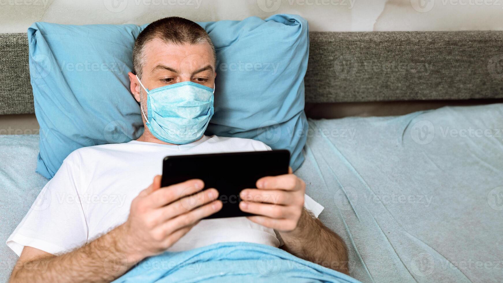 Coronavirus Quarantine. Middle-Aged Man In Mask Using Digital Tablet Watching Movie Lying In Bed At Home. Selective Focus photo