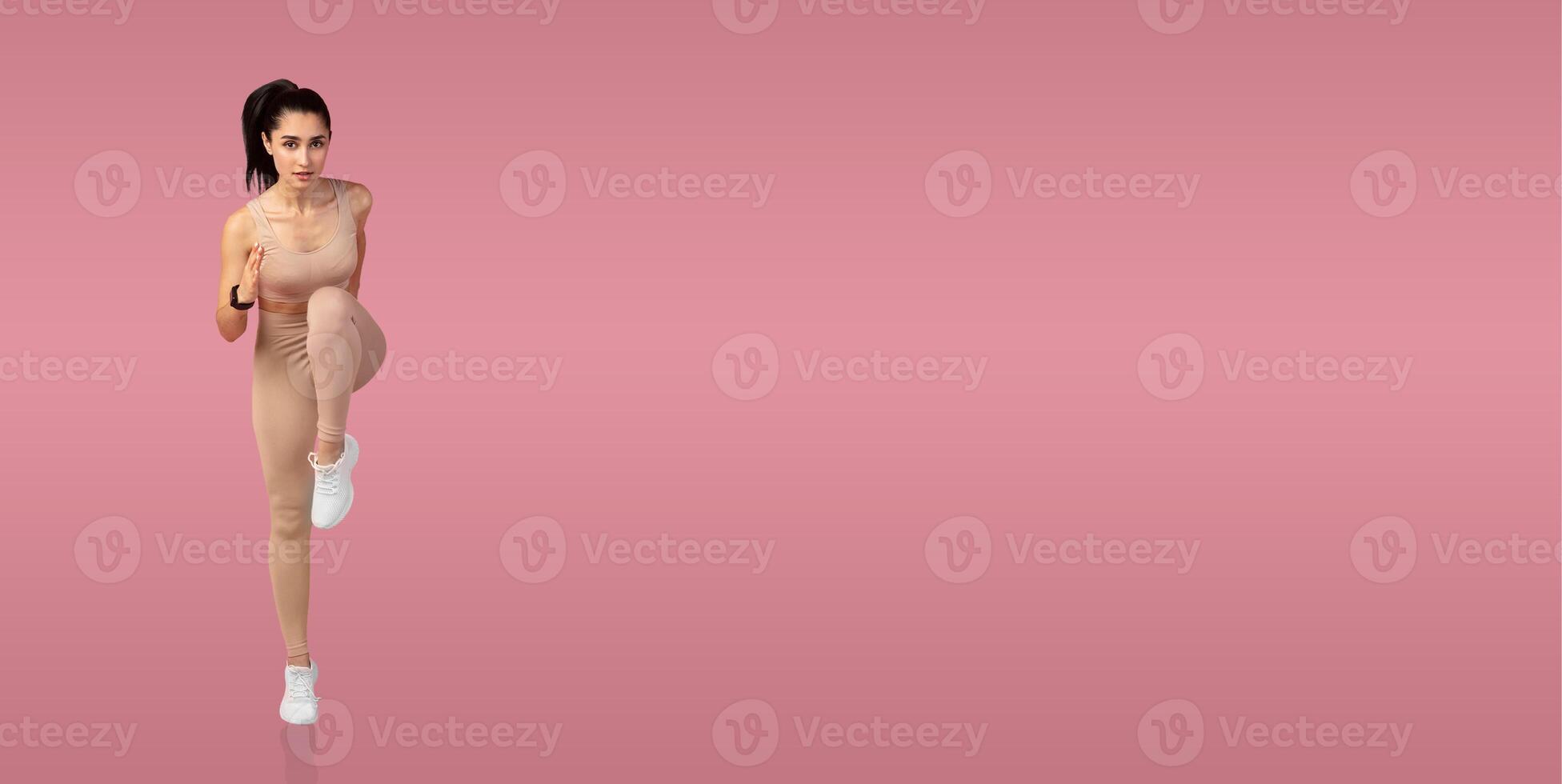 A young woman performs a knee lift exercise, showcasing her fitness routine in a bright pink studio setting. She is focused and dressed in comfortable workout attire. photo