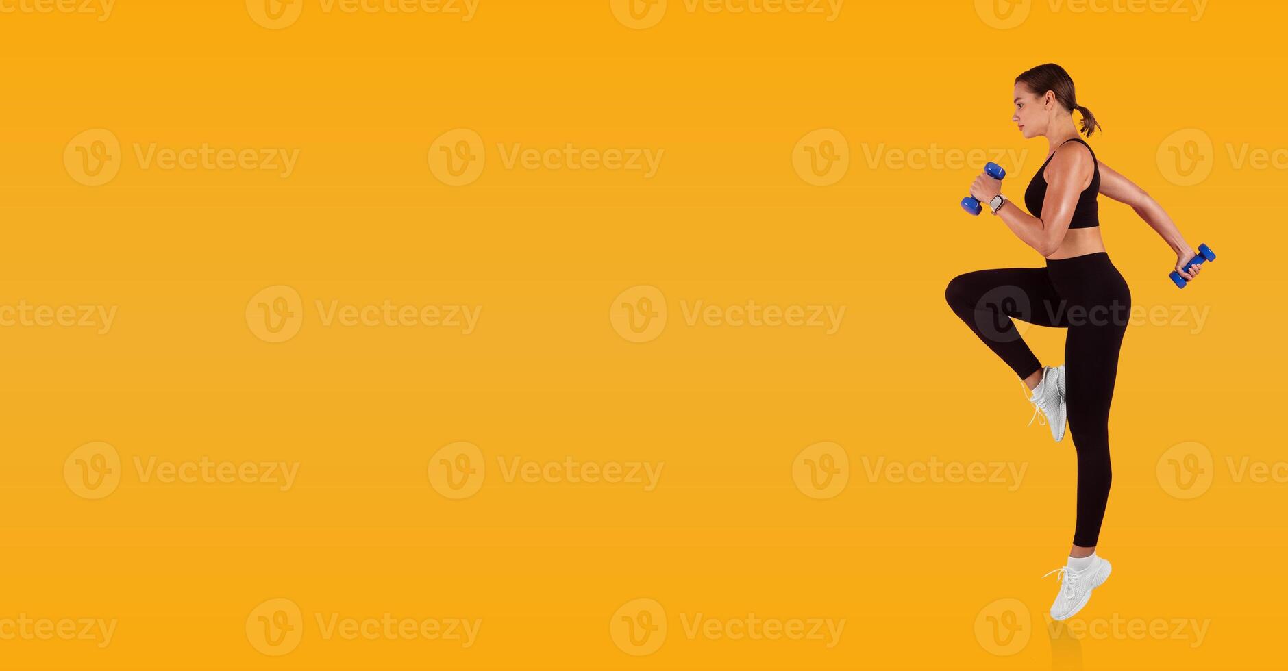 A woman is engaged in a dynamic workout, lifting two dumbbells while performing a high knee exercise. She is dressed in athletic wear, showcasing her fitness routine against a bright yellow backdrop. photo