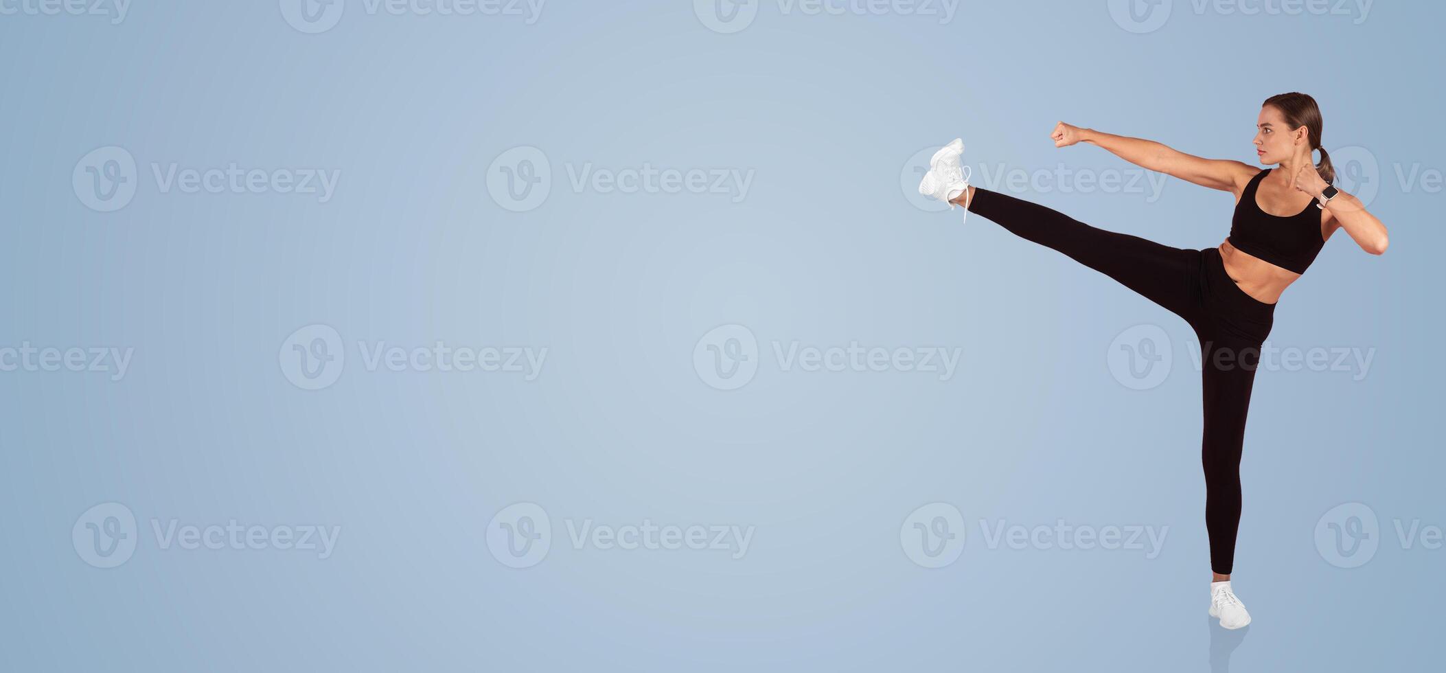 A woman in black athletic clothing executes a high kick with precision. The backdrop is a calm blue, emphasizing her movement and focus on fitness and agility. photo