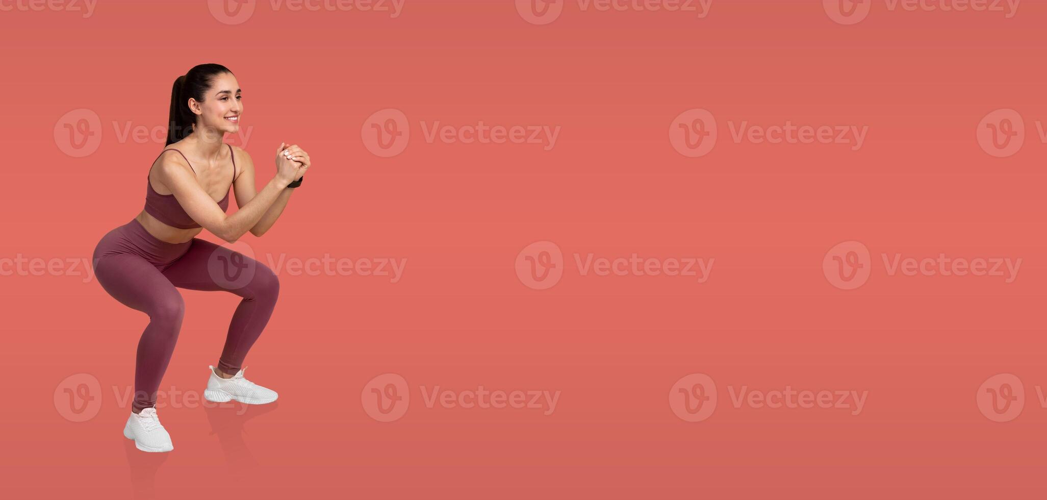 A young woman in athletic wear is performing squats with proper form against a vibrant background. Her focus and technique highlight dedication to fitness and strength training. photo