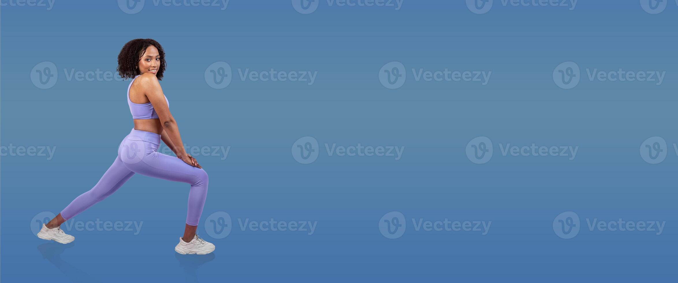 A woman wearing a lavender athletic set performs a stretching exercise, showcasing her form and flexibility. The clean blue background enhances the focus on her movement. photo