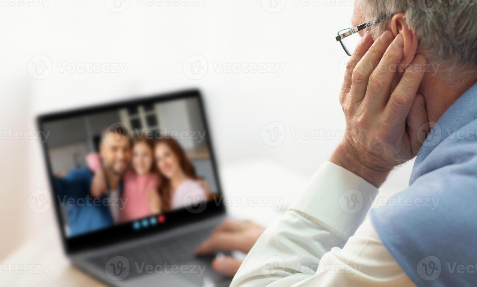 An elderly man is participating in a call with his family members, who appear joyful on the screen. He sits comfortably at home, enjoying the interaction. photo