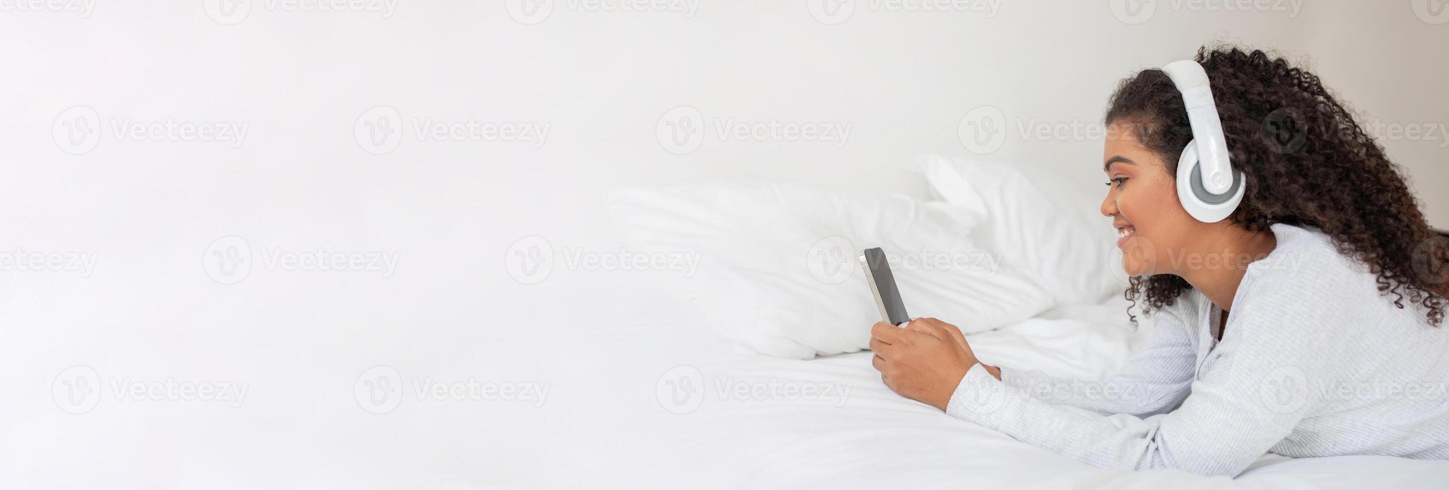 A young woman enjoys music through her headphones, relaxed on a bed surrounded by soft pillows. She is focused on her smartphone, engaging with her favorite tracks in a cozy setting. photo