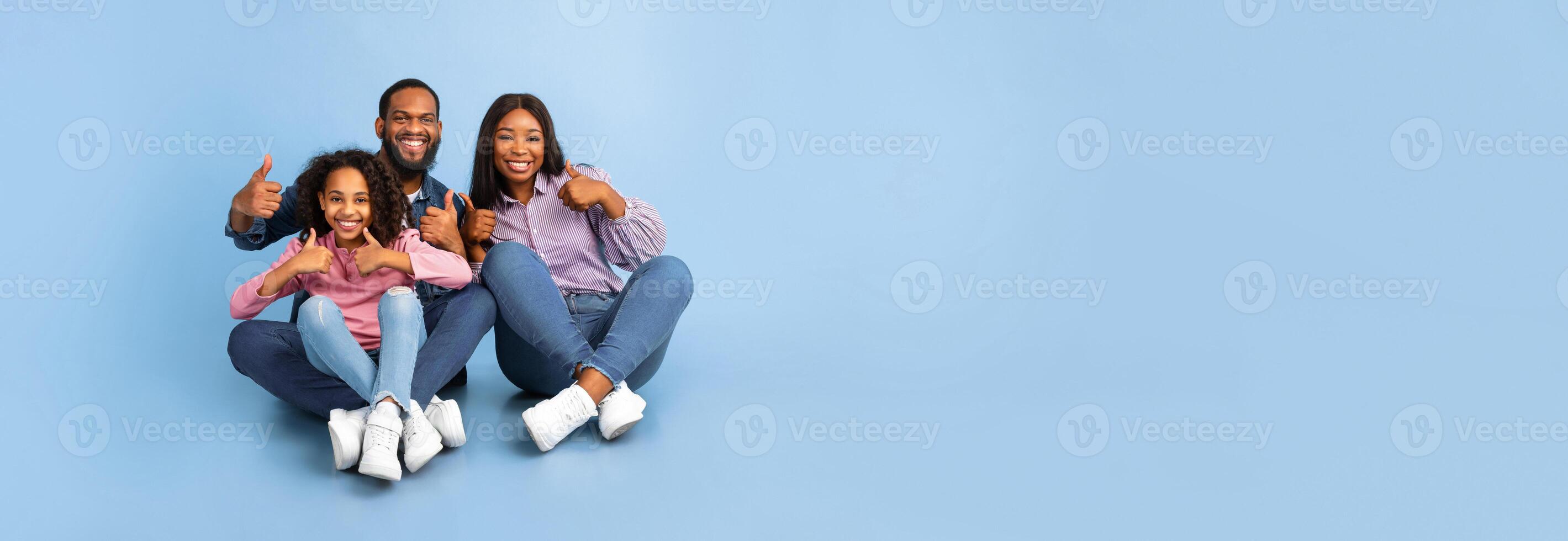 We Like It. Excited Black Mother, Father And Daughter Gesturing Thumbs Up Looking At Camera Sitting On The Floor Over Blue Background. Studio Shot. Panorama, Banner, Free Copy Space For Ad Or Design photo