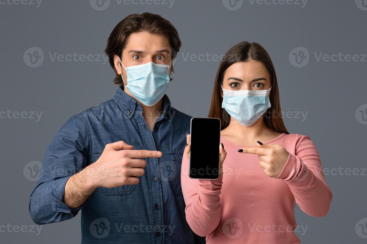 Coronavirus News Concept. Shocked Couple In Medical Masks Pointing At Smartphone With Blank Black Screen In Hands, Standing On Light Background photo