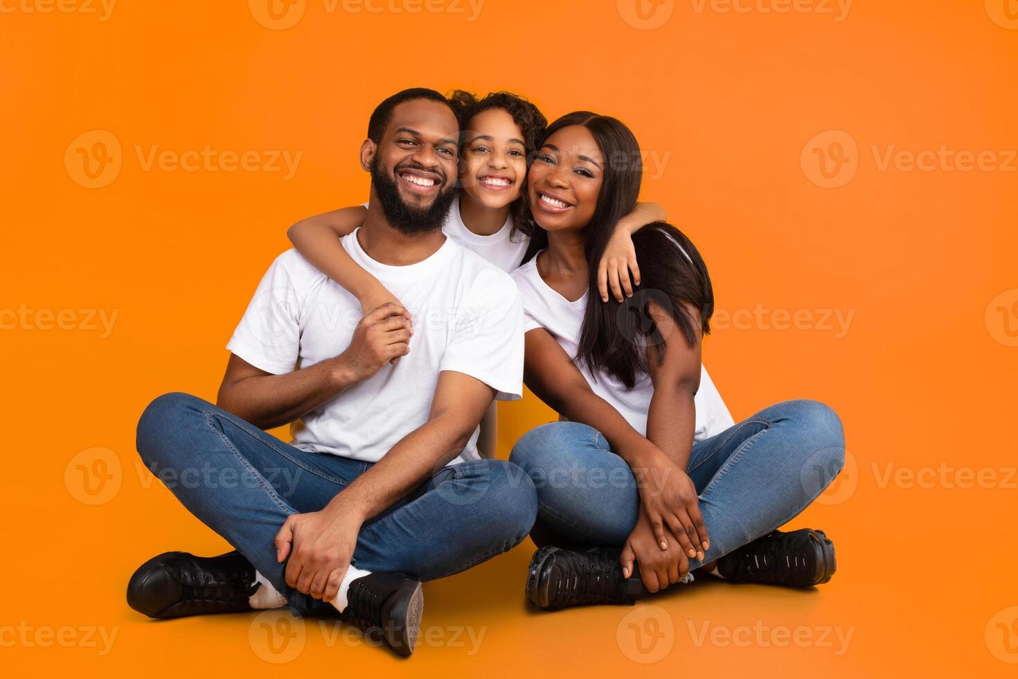 Happy African American man, woman and girl sitting on the floor isolated on yellow studio wall. Smiling daughter emracing her mother and father, looking at camera. Healthy Family Relationship Concept photo