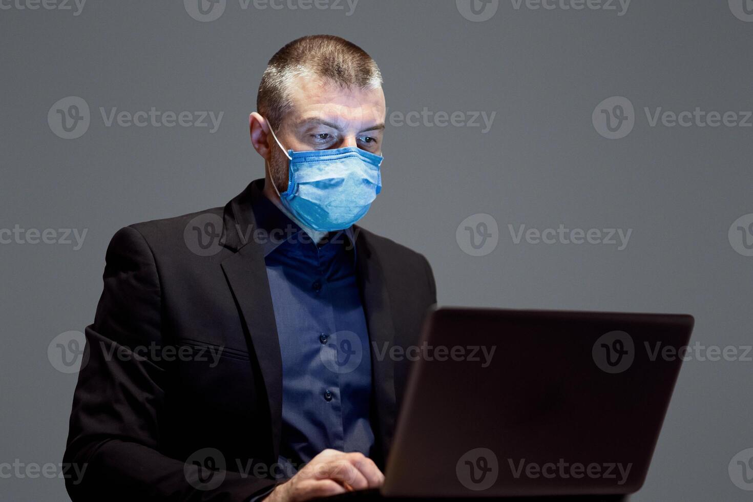 Businessman In Protective Medical Mask Working On Laptop Sitting Over Gray Studio Background. Work From Home. Selective Focus photo