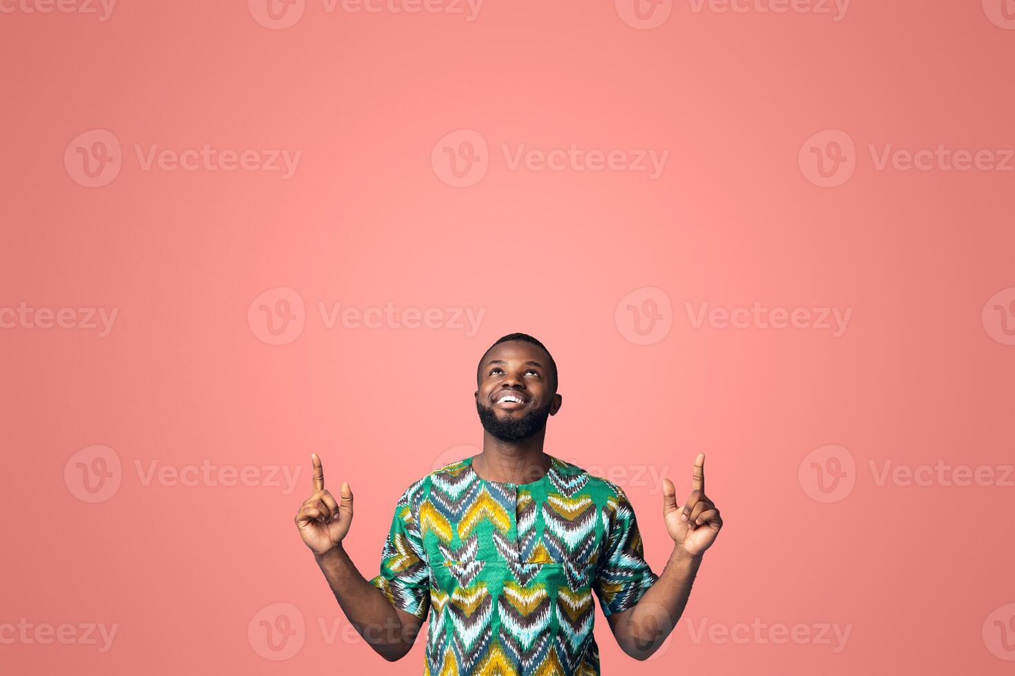 Happy black guy in african costume pointing up at free space, showing advertisement on blue studio background. Positive african american man looking up, directing at empty space for text photo