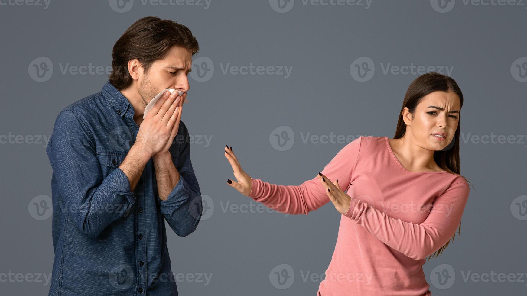 Young woman feeling disgust next to her sick male friend, trying to keep distance, standing together on grey background in studio, free space photo