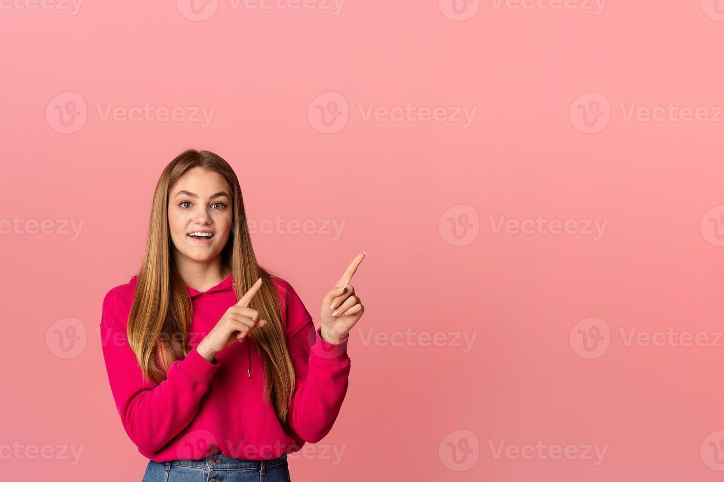 Funny smiling teenager girl with open mouth points with fingers towards an empty space, on pink background, studio shot photo