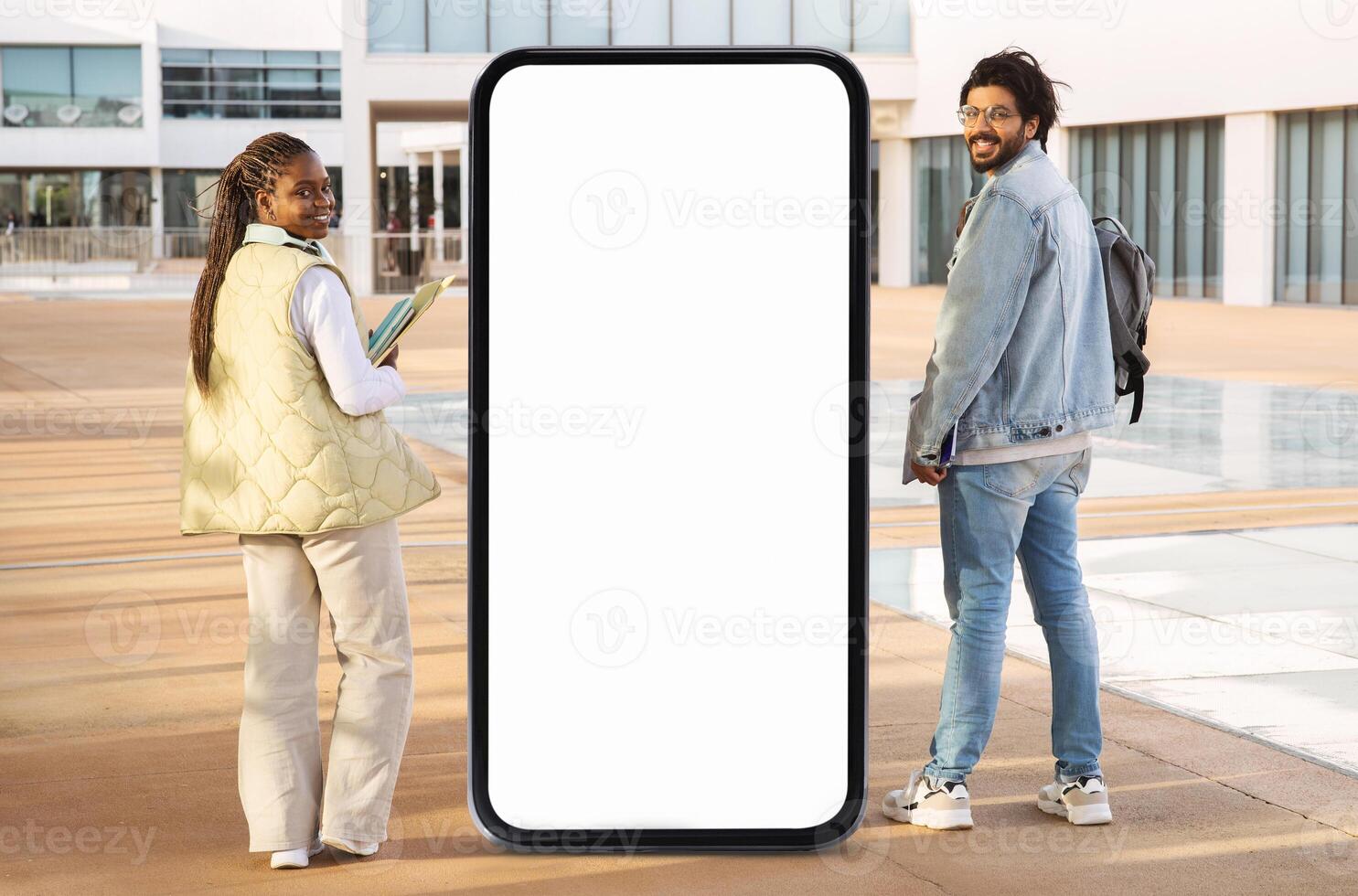 Two young adults pose outdoors beside a large blank screen, enjoying a sunny day. They are fashionably dressed, with one holding a notebook while smiling at the camera. photo