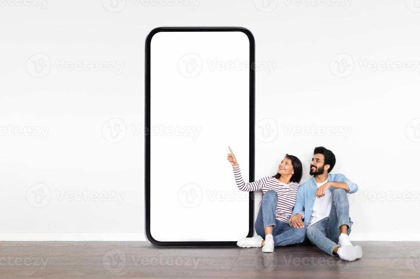 A young Indian couple sits on the floor, smiling and pointing at a large blank smartphone display. They are in a modern room with minimal decor, creating a relaxed atmosphere. photo