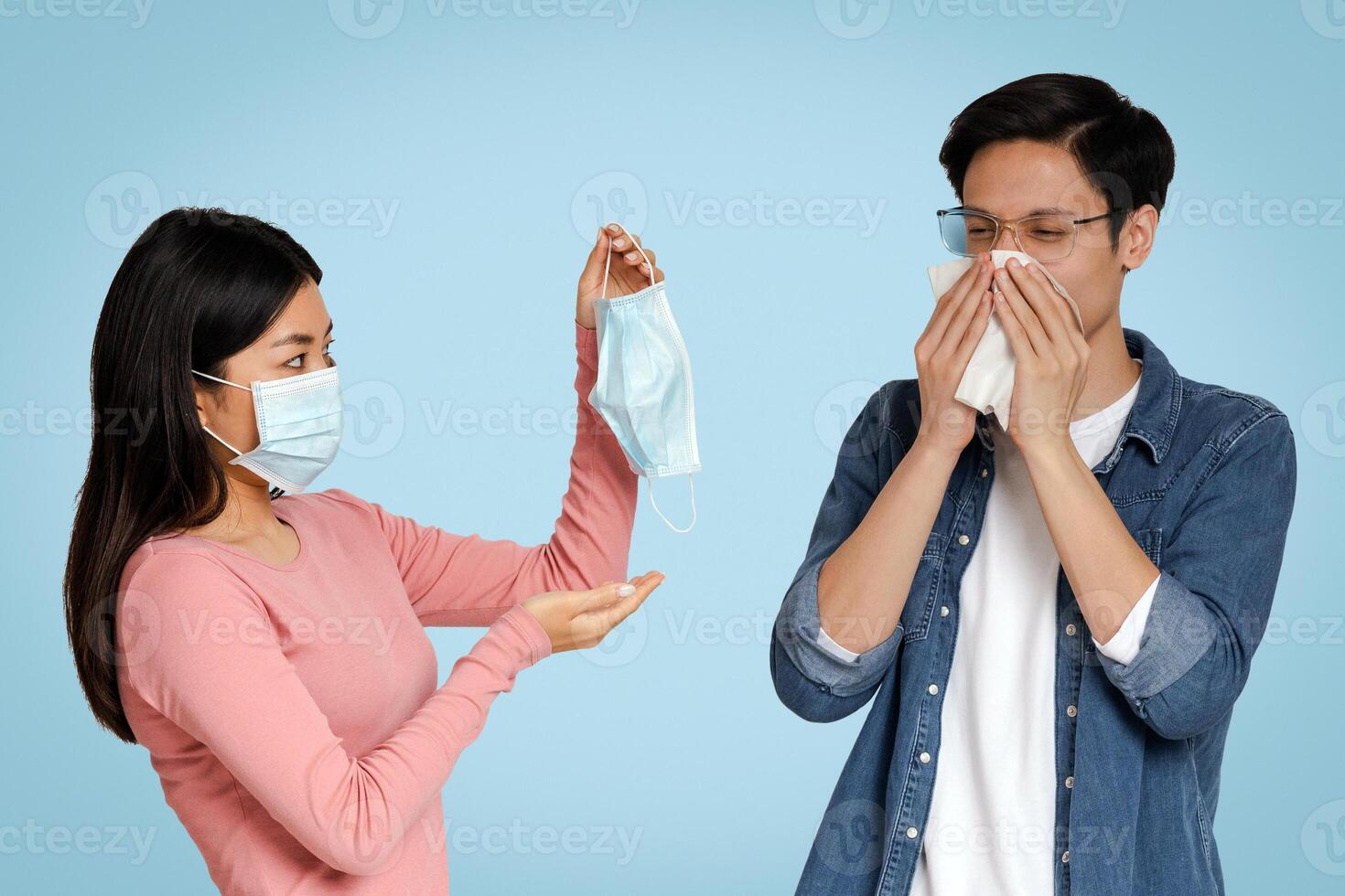 Virus Protection. Young Asian Woman Offering Medical Face Mask To Her Sick Boyfriend That Blowing The Nose Over Light Background, Free Space photo