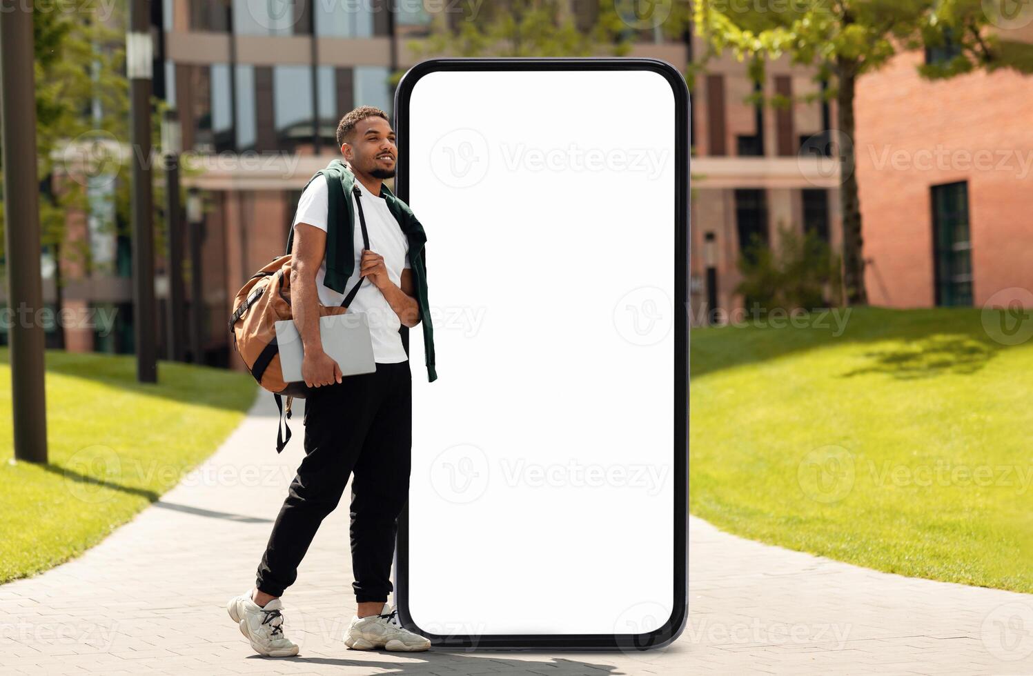 A young black man carrying a backpack leans against a large blank smartphone structure in a sunny outdoor setting. The area features green grass and modern buildings in the background. photo