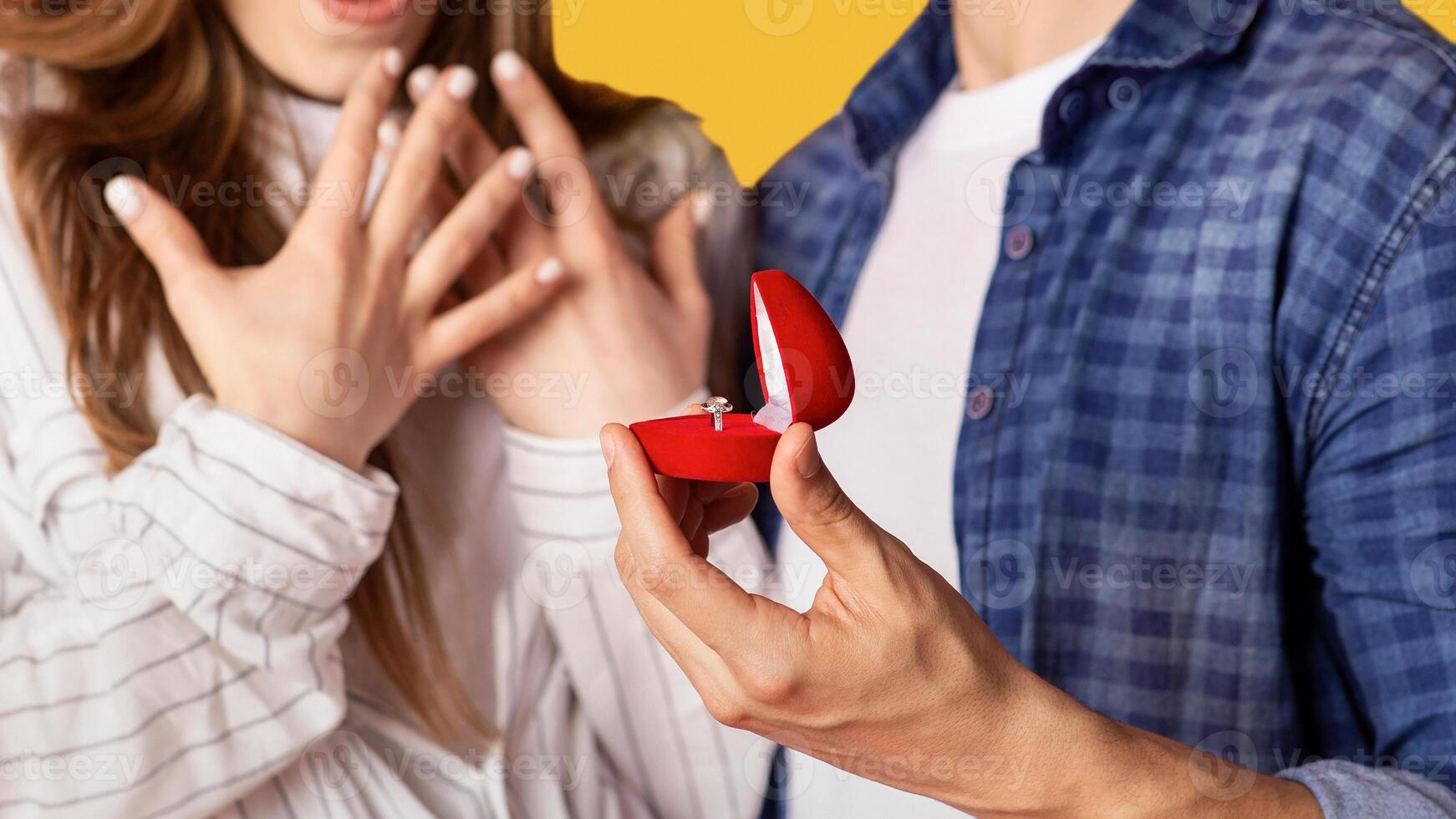 Marriage proposal and people emotions. Focus on box with ring in strong man hand and excited gestures of happy and surprised woman, isolated on yellow background, studio shot, close up, cropped photo