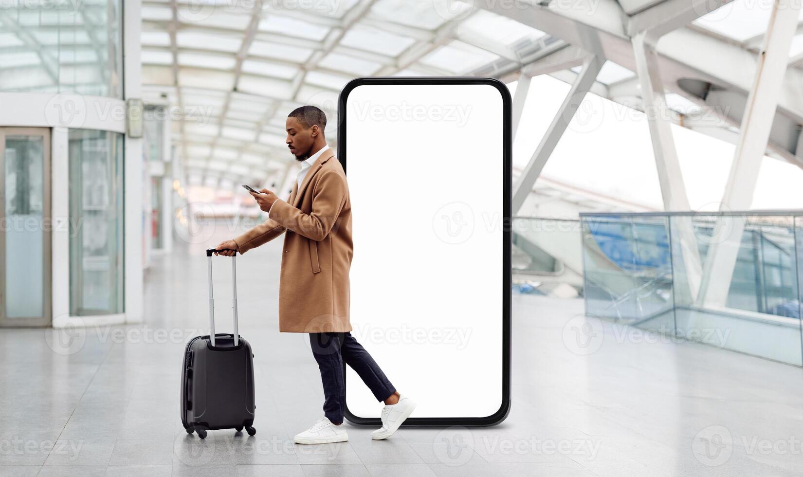 A traveler black guy stands in an airport terminal, pulling a suitcase while looking at his phone. A large blank screen looms in the background, adding to the contemporary atmosphere. photo