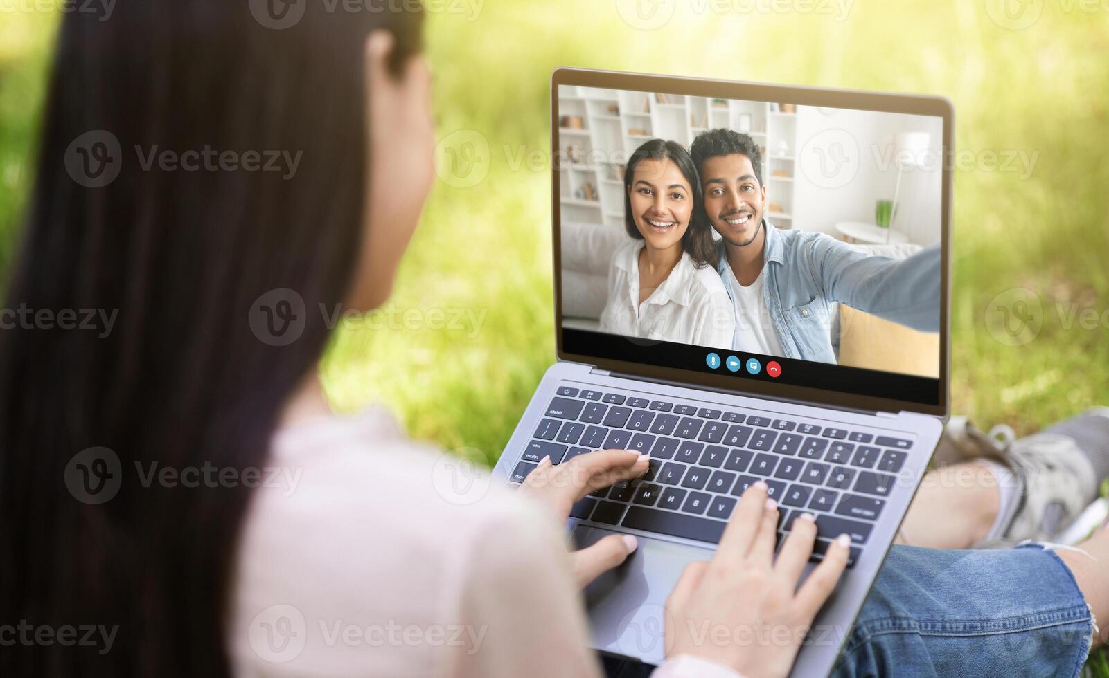 A woman sits on the grass, interacting with friends through a laptop. She smiles while they share moments during a virtual call in a sunny outdoor setting. photo