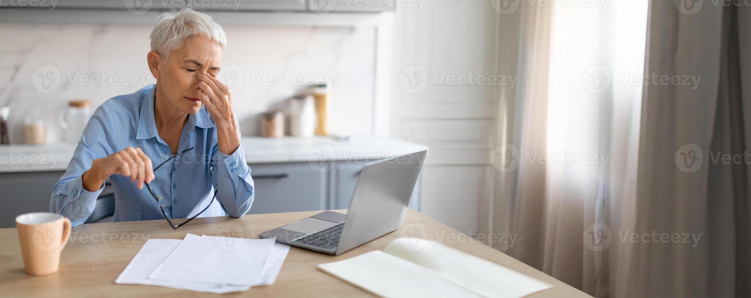 A senior woman sits at a kitchen table, rubbing her temples in frustration while looking at her laptop. Papers are scattered around as she deals with a challenging task. photo