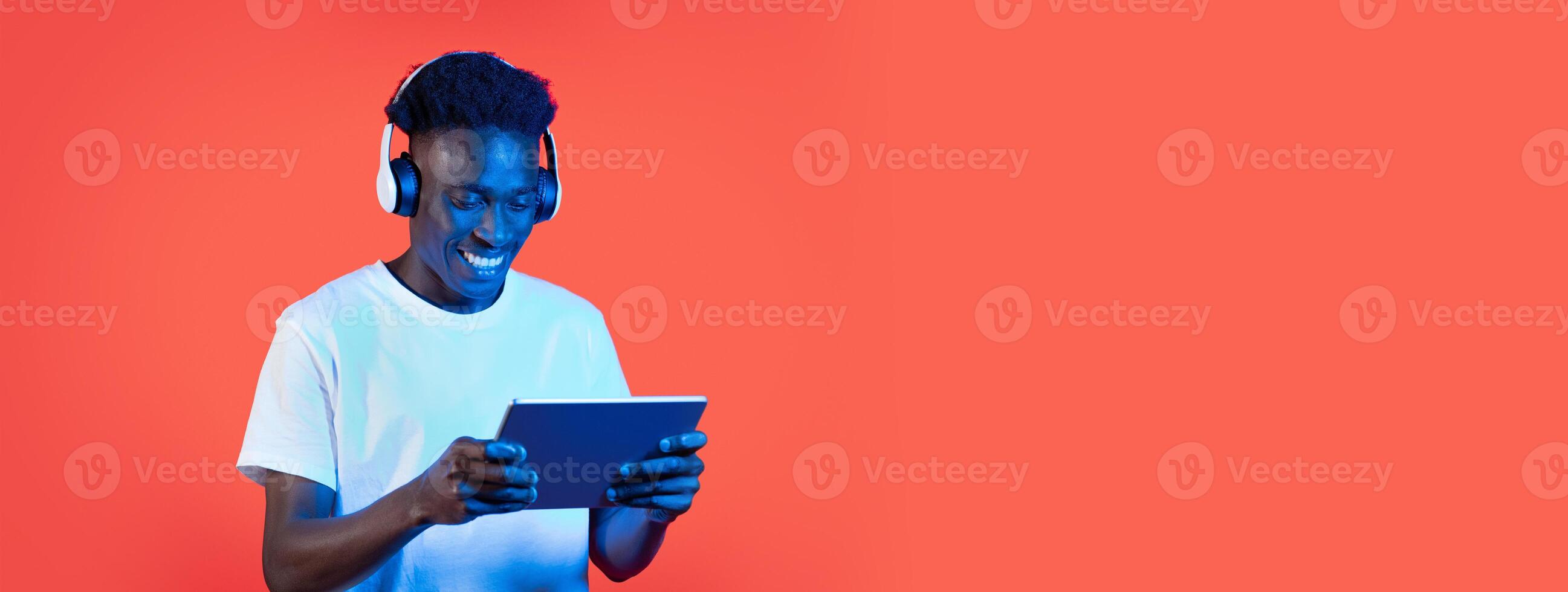 A cheerful young African American man is listening to music through headphones while playing on a tablet. The bright orange background adds a fun atmosphere to his engaging activity. photo