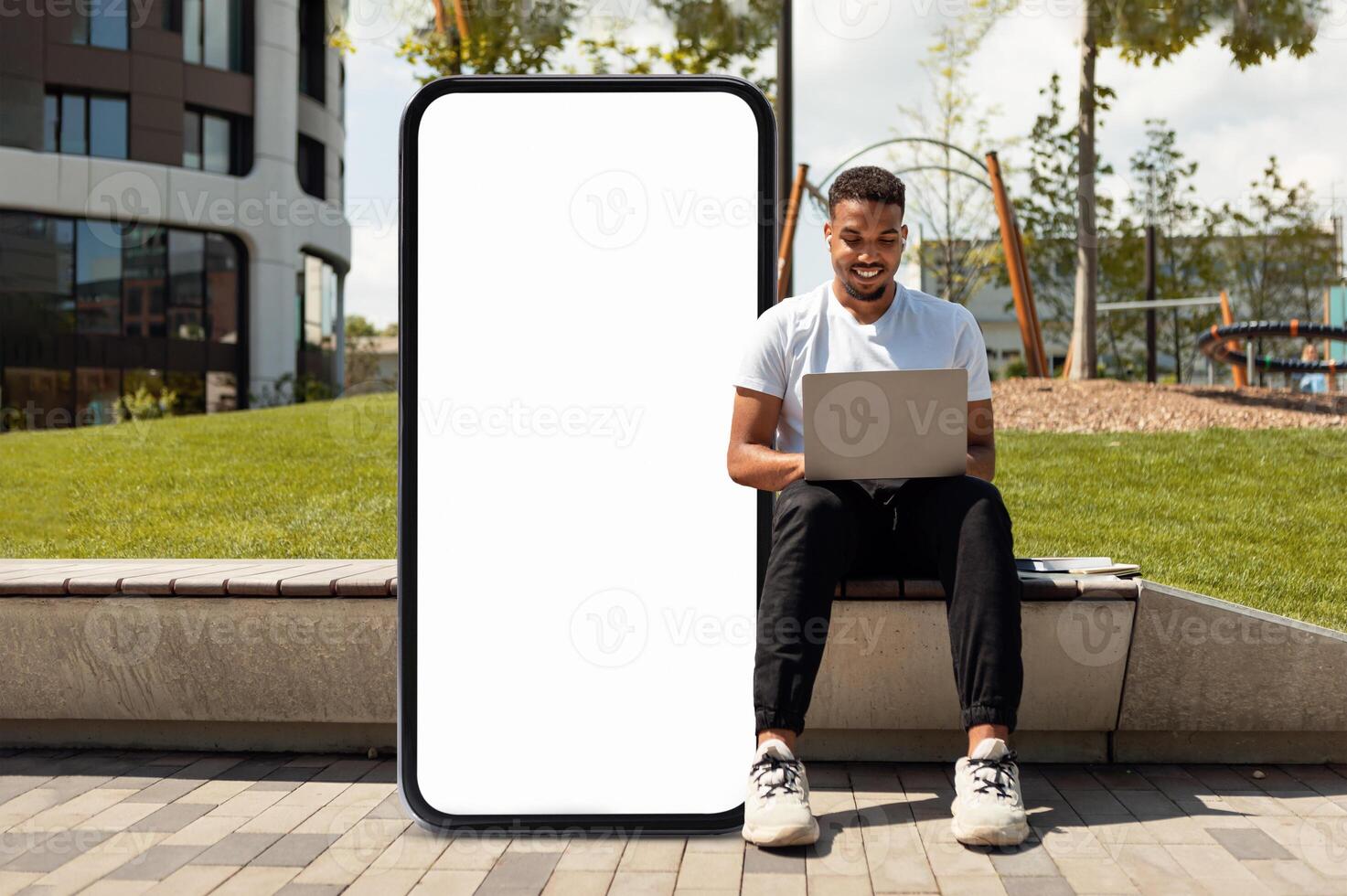 A young black man sits on a bench in a park, focused on his laptop while an enormous blank screen stands beside him, showcasing a relaxed atmosphere surrounded by greenery. photo