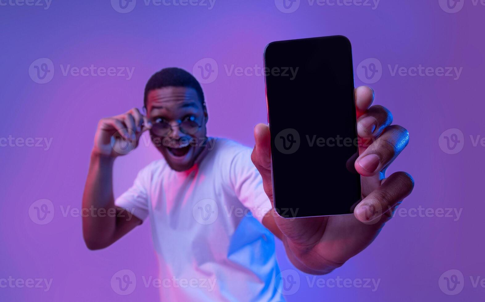 Cool Mobile App. Mockup Image Of Smartphone With Black Blank Screen In Hand Of Excited African American Guy In Sunglasses Standing In Vivid Neon Light Over Purple Studio Background, Copy Space photo