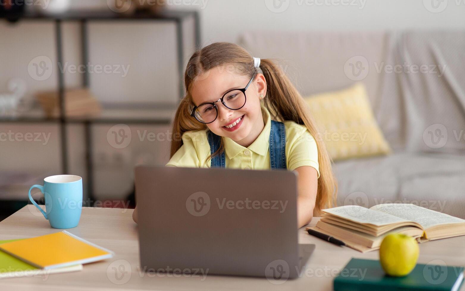 Distance Education. Smiling female pupil in eyeglasses sitting at desk, using laptop and typing on keyboard, doing homework, watching tutorial, lecture or webinar, studying online at home. E-learning photo