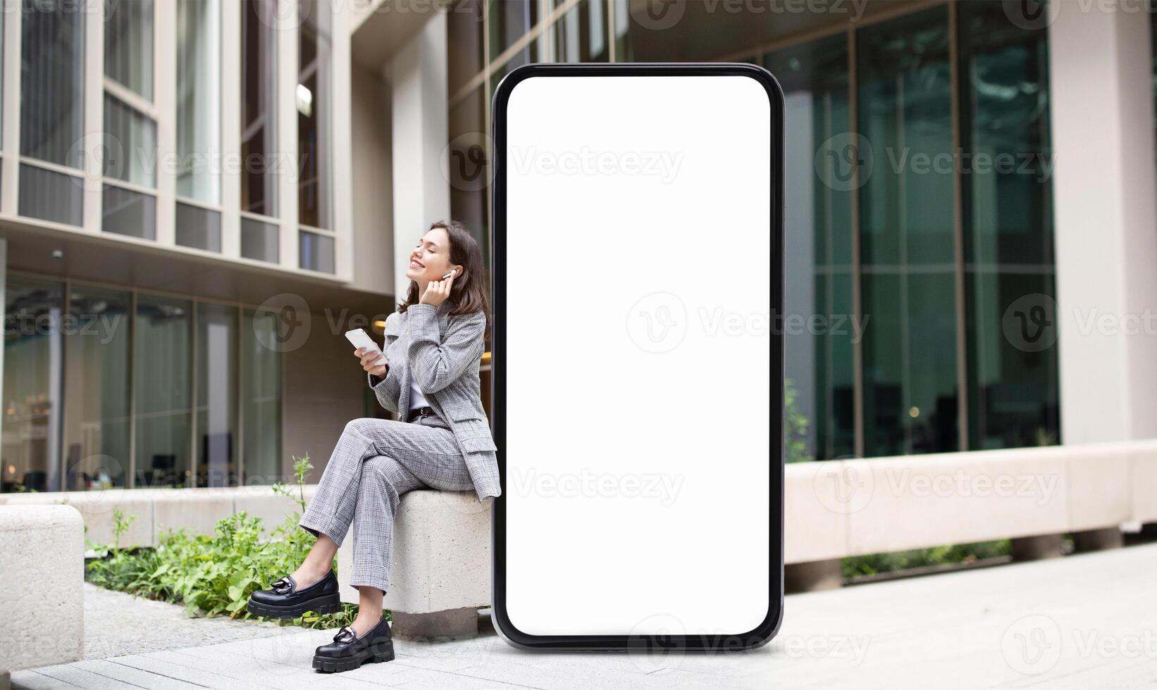 A woman in a business suit sits on a ledge by huge phone with blank screen outside a contemporary office building, engaged on her smartphone while holding a beverage. The atmosphere is calm and urban. photo